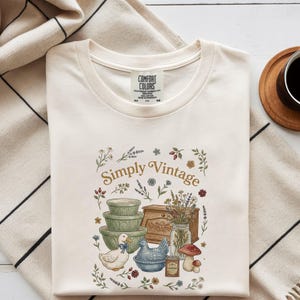 May include: Cream-colored t-shirt with a round neckline. The shirt features a "Simply Vintage" graphic with illustrations of bowls, a duck, a hen, flowers, and a mushroom. The shirt is made of a soft, comfortable material.