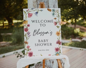 May include: A white sign with a floral border and the text "Welcome to Blossom's Baby Shower June 15th, 2024". The sign is on a wooden easel.