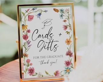 May include: A gold framed sign with a white background and a floral border. The sign says "Cards + Gifts for the Graduate thank you!" with a graduation cap illustration.