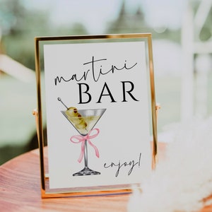 May include: A gold framed sign with the text "martini BAR enjoy!" in black script font. A watercolour illustration of a martini glass with olives and a pink ribbon is in the centre of the sign.