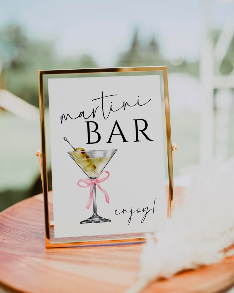 May include: A gold framed sign with the text "martini BAR enjoy!" in black script font. A watercolour illustration of a martini glass with olives and a pink ribbon is in the centre of the sign.