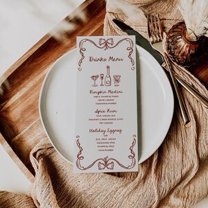 May include: A white card with a red border and a drinks menu for a holiday party. The menu includes Pumpkin Martini, Spice Rum, and Holiday Eggnog. The card is sitting on a white plate with a brown napkin and gold cutlery.