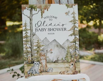 May include: A wooden easel displays a watercolor baby shower sign. The sign reads "Welcome to Olivia's Baby Shower" with a forest scene featuring mountains, trees, and woodland animals. The date 10.22.26 is also included.