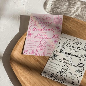Puede incluir: Dos invitaciones a una fiesta de graduaci&oacute;n con dibujos a tinta negra y rosa de un graduado con birrete y toga, un diploma y otras im&aacute;genes con tem&aacute;tica de graduaci&oacute;n. Las invitaciones dicen "Cheers to the Graduate" e incluyen el nombre "Olivia Richardson". Las invitaciones est&aacute;n sobre una superficie de madera.