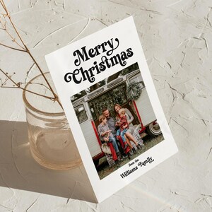 May include: A white Christmas card with the text "Merry Christmas" in a retro font. The card features a family photo in front of a vintage camper. The text "from the Williams Family" is printed below the photo.