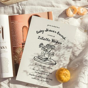 May include: A black and white illustration of a baby shower brunch invitation with a floral arrangement on a table. The invitation includes the text "Baby Shower Brunch", "in honor of", "Liliette Parker", "Ten O'Clock in the morning", "05.10.15", "The Ivy, 13 N Robertson Blvd", "Los Angeles, CA 90048", "Registered at Pottery Barn Baby", and "RSVP to Claire at 000.000.0000".