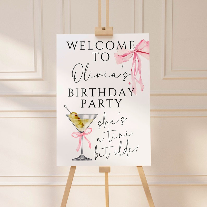 May include: A white sign with black text that reads "Welcome to Olivia's Birthday Party, she's a  tini bit older". A pink ribbon with a bow is in the top right corner. A martini glass with olives and a pink ribbon is in the bottom left corner.