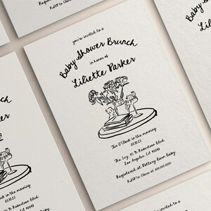 May include: A black and white illustration of a baby shower brunch invitation with the text "Baby Shower Brunch" and "Liliette Parker". The illustration includes a tray with a vase of flowers, books, and a glass.
