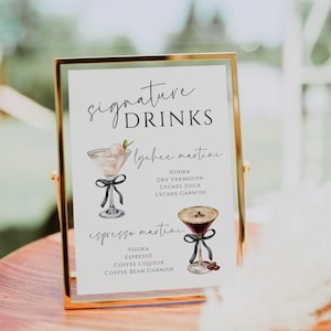 May include: A gold-framed sign with the text "signature DRINKS" and two illustrated cocktails. The first cocktail is a lychee martini with the ingredients listed below: vodka, dry vermouth, lychee juice, and lychee garnish. The second cocktail is an espresso martini with the ingredients listed below: vodka, espresso, coffee liqueur, and coffee bean garnish.