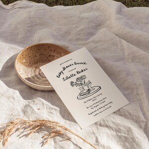 May include: A black and white illustration of a baby shower brunch invitation for Liliette Parker. The invitation includes the date, time, and location of the event. The illustration features a bouquet of flowers, a book, and a cup.