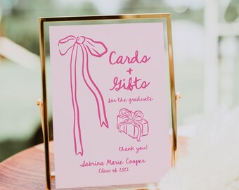 Op de afbeelding: Een gouden lijst met een roze achtergrond en roze tekst die "Kaarten + Cadeaus voor de afgestudeerde bedankt! Sabrina Marie Cooper Klas van 2015" luidt. Het bord heeft een roze lint en een roze cadeaudoos.