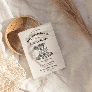 May include: A white card with black text and a hand-drawn illustration of flowers in a vase. The text reads "You're invited to a Baby Shower Brunch in honor of Liliette Parker. The Olive & The Oak, 10:00 in the morning, The Story, 55 N. Robertson Blvd, Los Angeles, CA 90048. RSVP to Pottery Barn Baby, KJP@OliveandtheOak.com"