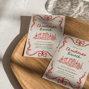 May include: Two Christmas Brunch invitations on a wooden tray. The invitations have a red and green design with the words "Christmas Brunch" and event details. The invitations are decorated with festive illustrations.