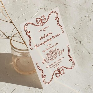 May include: A white invitation card with a red border and a red bow at the top. The text on the card reads "You're invited to the Hudson's Thanksgiving Dinner 4 pm 18th November 2014 1145 Birk Share Dr, Gloucester, MA RSVP to Kate by Nov 23.458.209".
