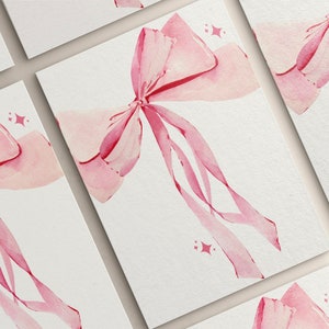 May include: A set of five blank cards featuring a watercolour illustration of a pink bow on each card. The bow is tied with a ribbon and has a subtle sparkle effect.