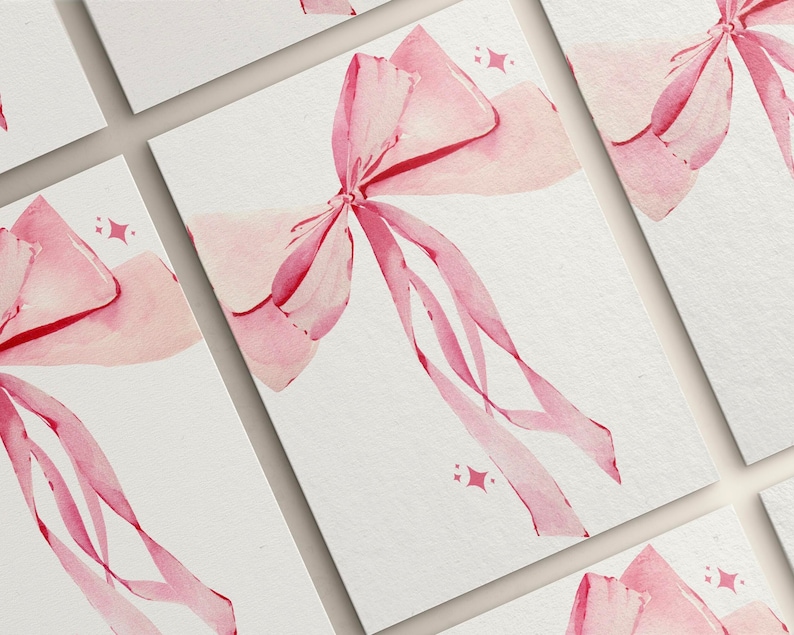 May include: A set of five blank cards featuring a watercolour illustration of a pink bow on each card. The bow is tied with a ribbon and has a subtle sparkle effect.