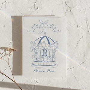 May include: A blue and white illustration of a carousel with a ribbon and bow at the top. The text "Olivia Rose" is printed below the carousel.