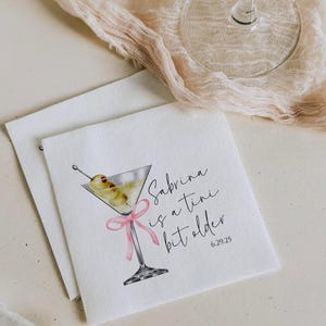 May include: White cocktail napkin with a watercolour illustration of a martini glass with olives and a pink ribbon. The text reads "Sabrina is a tiny bit older 6.29.25".