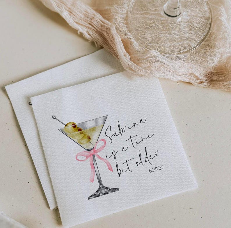 May include: White cocktail napkin with a watercolour illustration of a martini glass with olives and a pink ribbon. The text reads "Sabrina is a tiny bit older 6.29.25".