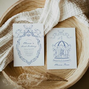 May include: Two blue and white invitation cards for a first birthday party. The first card features a decorative frame with the text "Olivia's First Birthday" and the second card features a carousel with the text "Olivia Rose".