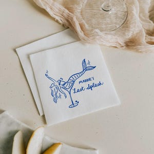 Può includere: Un tovagliolo di carta bianco con un'illustrazione blu di una sirena che tiene un bicchiere da cocktail. Il testo "Maddie's Last Splash" è stampato sotto l'illustrazione.