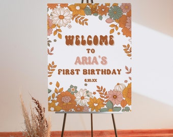 May include: Welcome to Aria's First Birthday sign with a white background and a floral border in shades of orange, yellow, green, and pink. The text is in a retro font style.