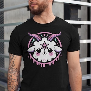 May include: Black t-shirt with a graphic of a cute, white, fluffy lamb with pink horns and a pentagram on its head. The lamb is surrounded by a pink circle with dripping pink paint.