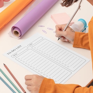 May include: A person is writing on a white to-do list with a pencil. The list has sections for name and date. There are colored pencils and a sticky note on the table.