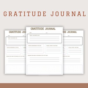 May include: Three white gratitude journals with lined pages and prompts for daily reflection. The words "Gratitude Journal" are printed at the top of each page and in a brown font above the journals. The background is a light beige color.