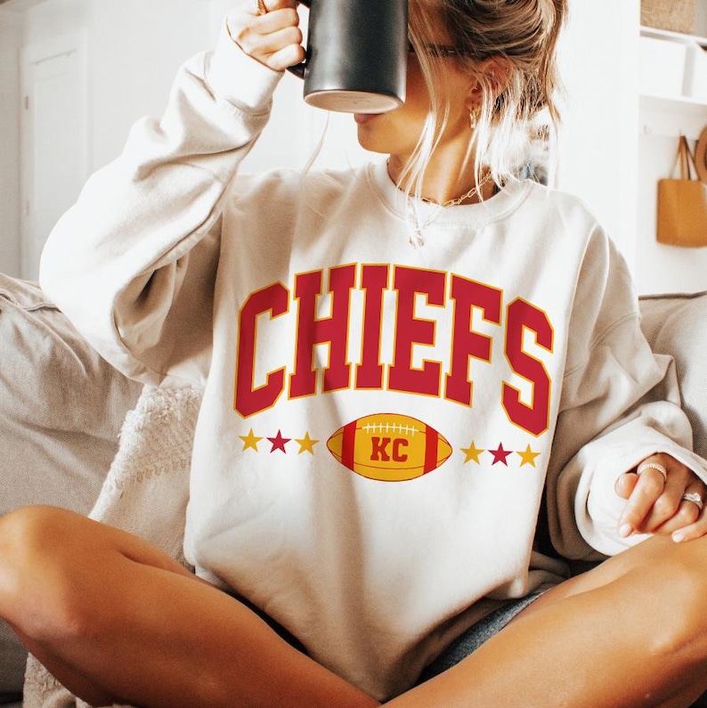 May include: Cream-coloured sweatshirt featuring the word "CHIEFS" in red block letters. Below the word is a yellow and red football graphic with "KC". Gold stars are on either side of the football.