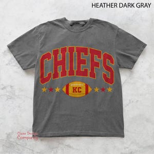 May include: Dark grey t-shirt featuring the word "CHIEFS" in red block letters with a gold outline. Below the word "CHIEFS" is a football with the letters "KC" and gold stars on each side. The text "Class Design Company" is in the bottom left corner.
