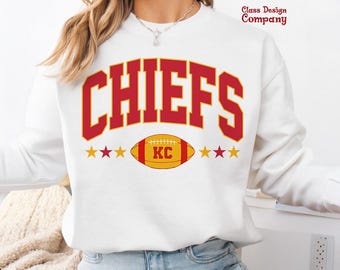 Puede incluir: Sudadera blanca con la palabra "CHIEFS" en letras rojas con contorno dorado. Debajo, un balón de fútbol dorado y rojo con "KC" en el centro. La sudadera también tiene estrellas doradas a cada lado del balón. El texto "Class Design Company" está en la esquina superior derecha.