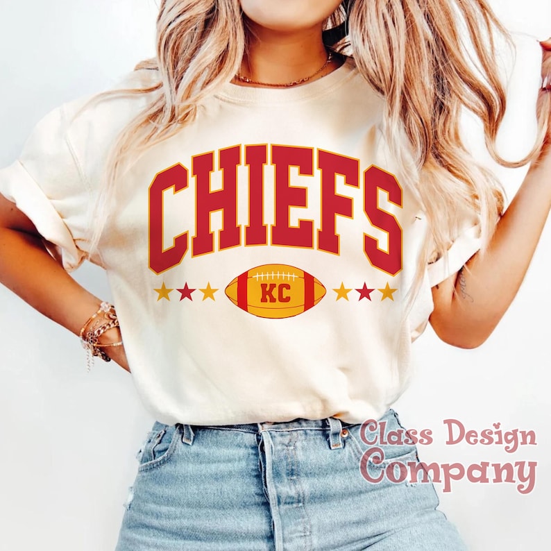 May include: Cream-coloured t-shirt featuring the word "CHIEFS" in red block letters. Below the word is a football with "KC" inside, with gold stars on either side. The shirt is a casual style.