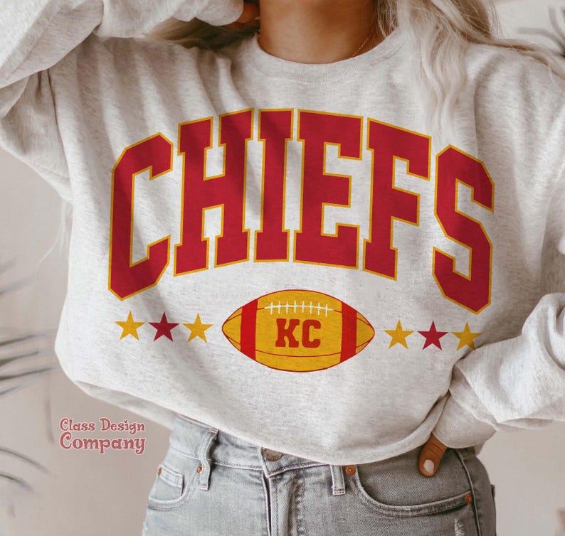 May include: A light grey sweatshirt featuring the word "CHIEFS" in red, bold lettering. Below is a yellow and red American football with "KC" in the centre. Three yellow stars flank the football on each side.