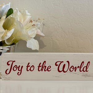 May include: A decorative wooden sign with the phrase "Joy to the World" in red script. The sign is white with a distressed finish. A silver vase with white flowers is to the left of the sign.