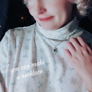 May include: A silver necklace with a small, square pendant. The pendant has a dark green center and a silver border. The text "you can make a necklace" is written on the shirt.
