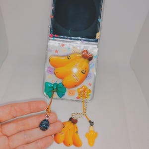 May include: Clear phone case with a banana design. The case features a yellow banana charm, a green bow, and a small blue charm. The phone case is decorated with small bear and star shapes.