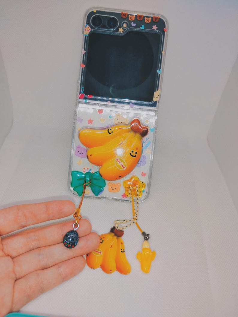 May include: Clear phone case with a banana design. The case features a yellow banana charm, a green bow, and a small blue charm. The phone case is decorated with small bear and star shapes.