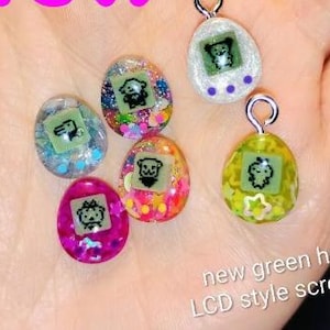 May include: A collection of colorful, egg-shaped pendants with silver loops. Each pendant features a small, green LCD-style screen with a cartoon character. The pendants are filled with glitter and have the text "new green hue LCD style screens".