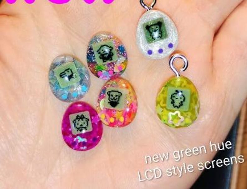 May include: A collection of colorful, egg-shaped pendants with silver loops. Each pendant features a small, green LCD-style screen with a cartoon character. The pendants are filled with glitter and have the text "new green hue LCD style screens".