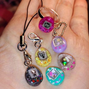 May include: A selection of small, colourful digital pet toys in various shapes and colours, including purple, yellow, and teal. Some have glitter and small charms. One is attached to a black lanyard and another to a silver ring.