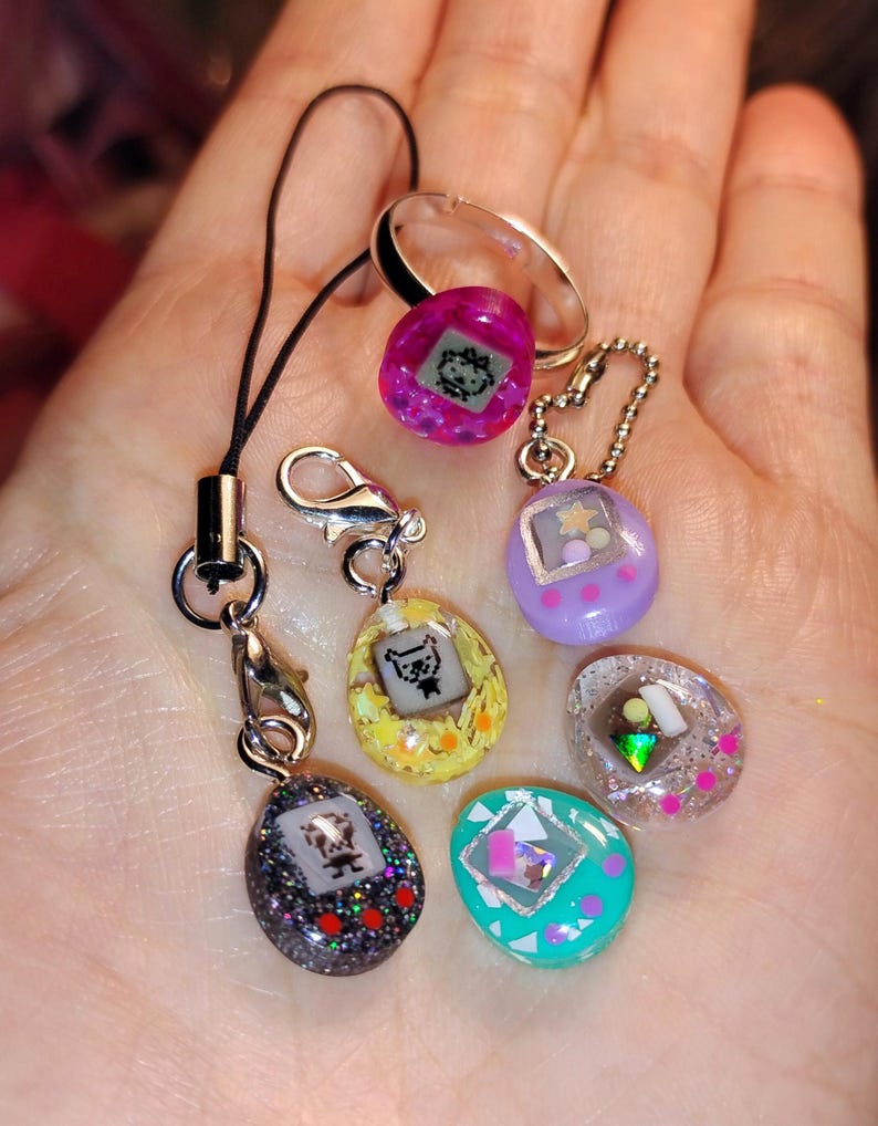 May include: A selection of small, colourful digital pet toys in various shapes and colours, including purple, yellow, and teal. Some have glitter and small charms. One is attached to a black lanyard and another to a silver ring.