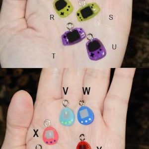 May include: A selection of miniature game console charms in various colours. The charms include grey, green, purple, pink, blue, and red. Each charm has a silver loop for attaching to jewellery or crafts. The charms are small, likely less than 2.5 cm in size.