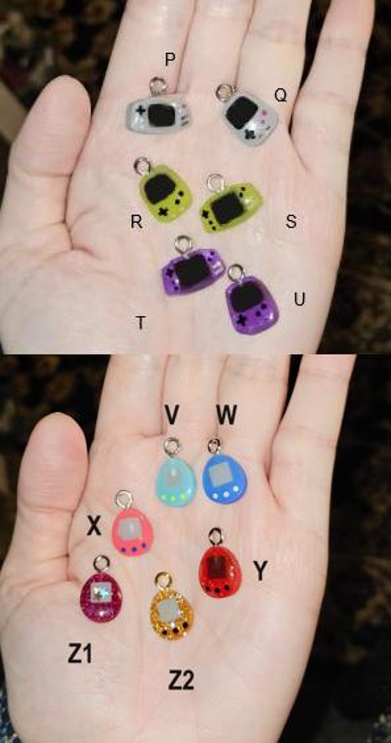 May include: A selection of miniature game console charms in various colours. The charms include grey, green, purple, pink, blue, and red. Each charm has a silver loop for attaching to jewellery or crafts. The charms are small, likely less than 2.5 cm in size.