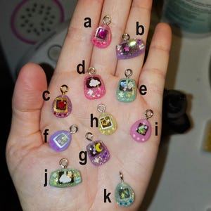 May include: A selection of small, colourful resin charms with gold-coloured loops. The charms feature various designs, including hearts, clouds, stars, and pixelated images. The charms are in shades of pink, purple, green, yellow, and blue, with glitter accents.