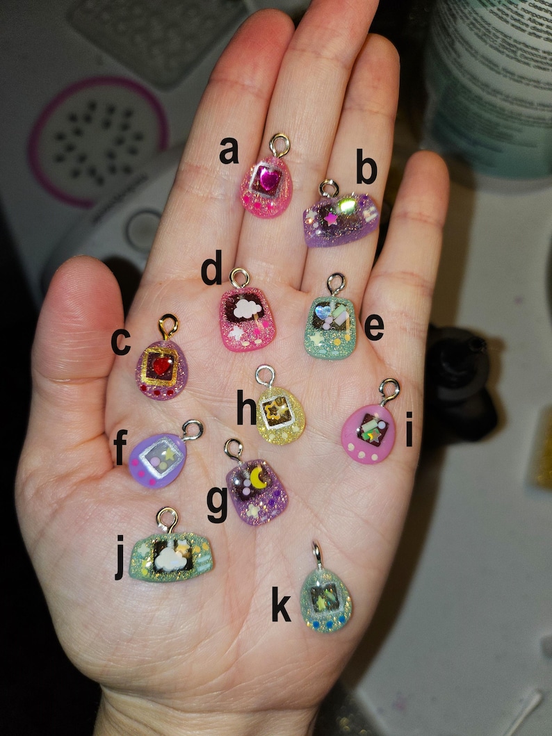May include: A selection of small, colourful resin charms with gold-coloured loops. The charms feature various designs, including hearts, clouds, stars, and pixelated images. The charms are in shades of pink, purple, green, yellow, and blue, with glitter accents.