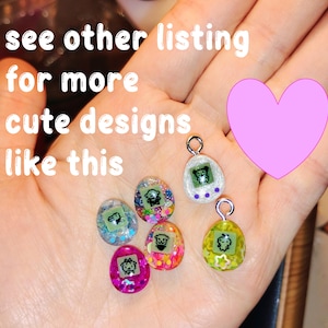 May include: A hand displaying six colourful, egg-shaped charms, each with a small cartoon character. The charms are in pink, green, and blue, with glitter and a silver loop. The image includes text: "see other listing for more cute designs like this."