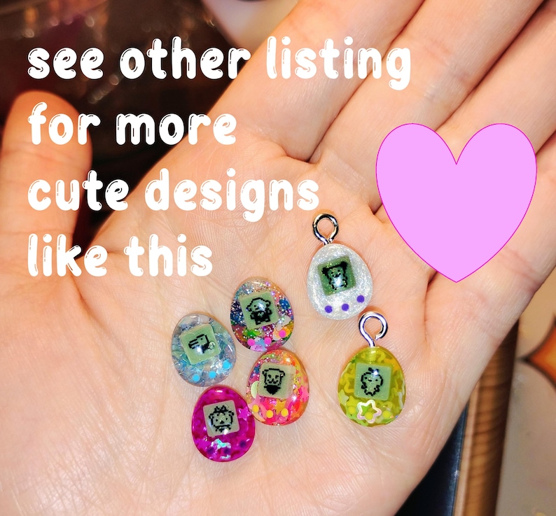 May include: A hand displaying six colourful, egg-shaped charms, each with a small cartoon character. The charms are in pink, green, and blue, with glitter and a silver loop. The image includes text: "see other listing for more cute designs like this."