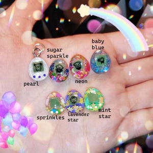 May include: A collection of seven colorful, miniature, egg-shaped pendants. Each pendant features a small screen displaying a pixelated character. The pendants are labeled with names like "pearl," "sugar sparkle," and "baby blue."