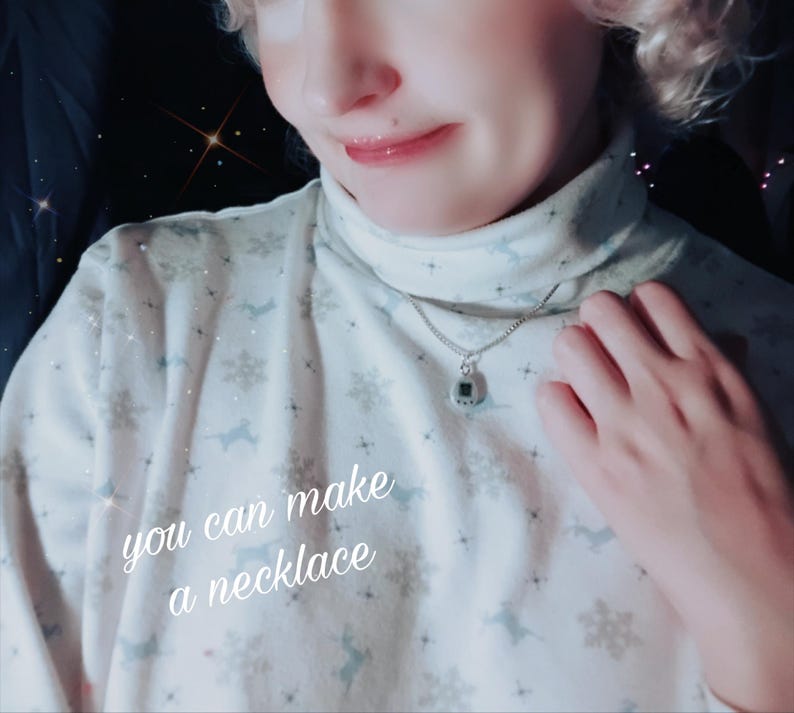 May include: A silver necklace with a small, square pendant. The pendant has a dark green centre and a silver border. The text "you can make a necklace" is written on the shirt.